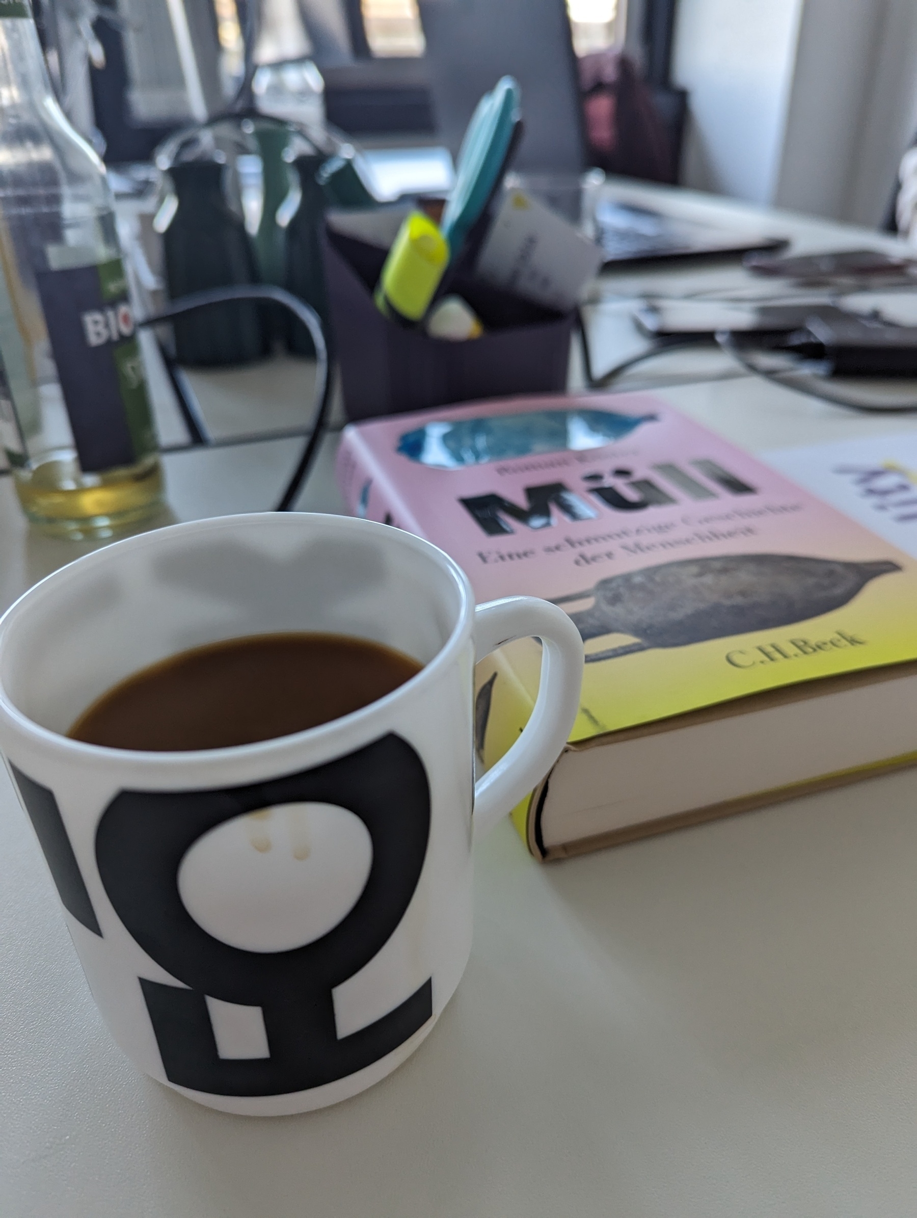 Snapshot from a desk. You see a black and white mug with coffee in there. Next to it is a colourful book cover boasting "Müll" aka waste.