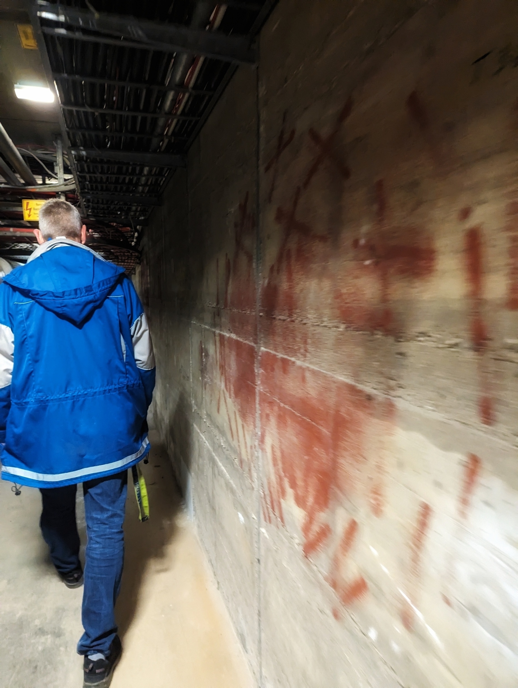 A man walks along a narrow concrete path. He wears blue maintenance staff clothing. He walks quickly. You see the concrete wall on the right with red paint fainting. Eerie