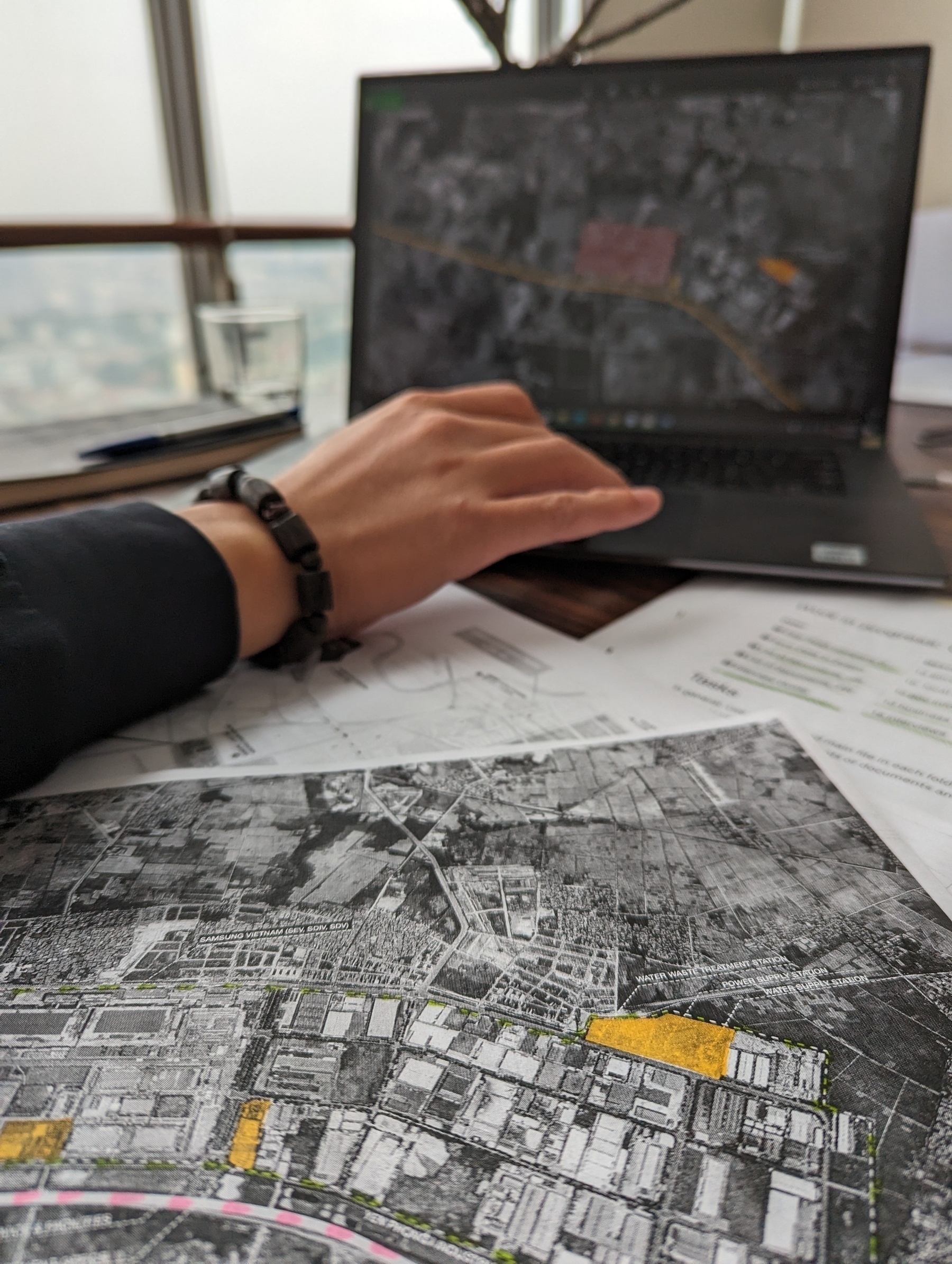 Maps on a table, printed and on a laptop. It's an aerial view of East Hanoi-ish. Some buildings are yellow. In the background, you see the actual Hanoi from above through a window 