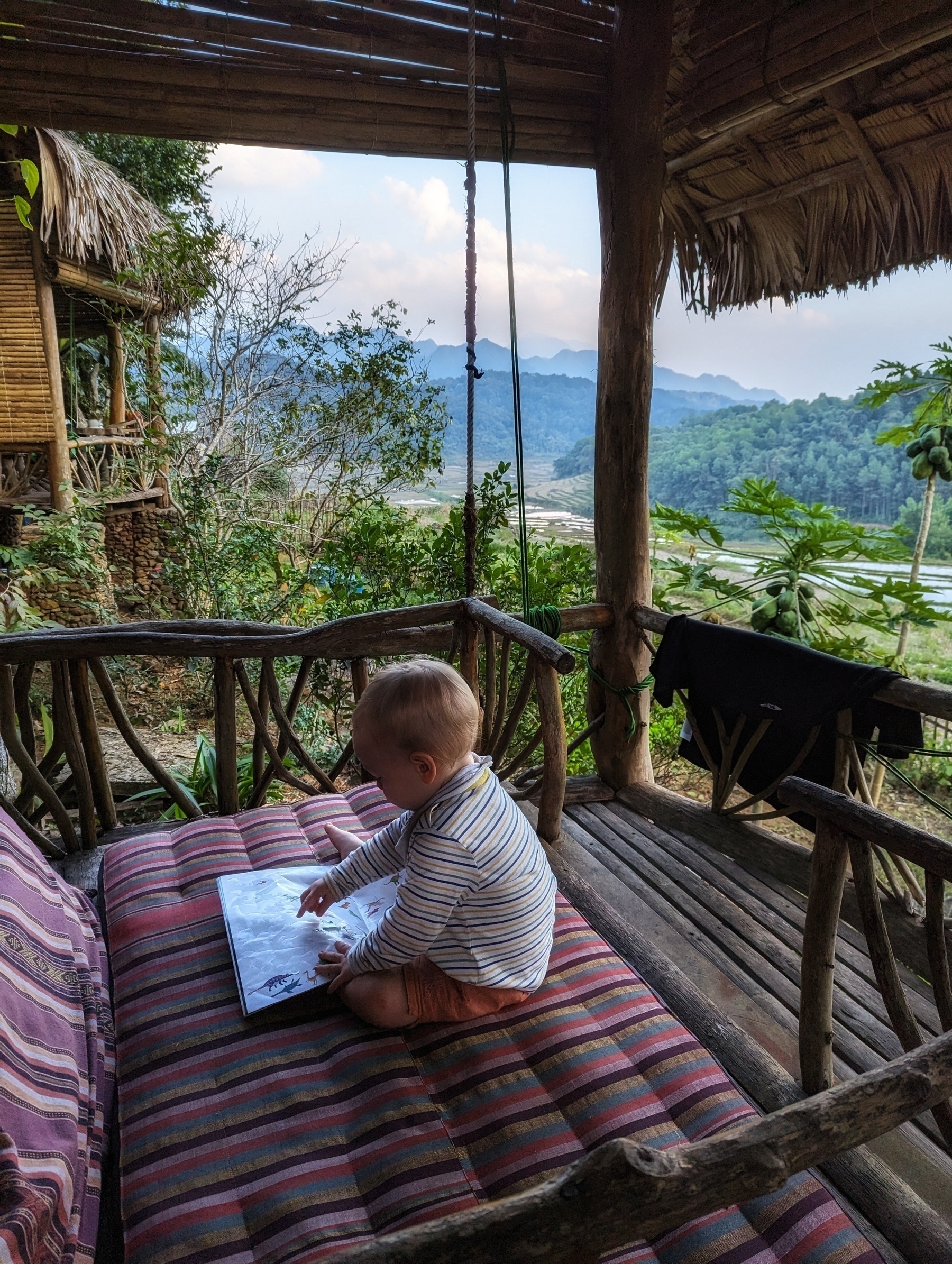 Picture of a little kid sitting on a wooden balcony playing with a sticker magazine. Huts on the left, scenic mountains and green rice fields in the back. A lot going on but still a very quiet snapshot.