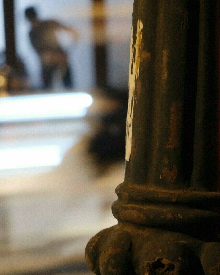Cropped photo shot from the curbside focusing on a rusty lamppost with a lady wiping the floor In the background. Sounds strange, but she's behind the window and taking care of the entrance. I think this is where they park their motorbikes, or just sit and chill
