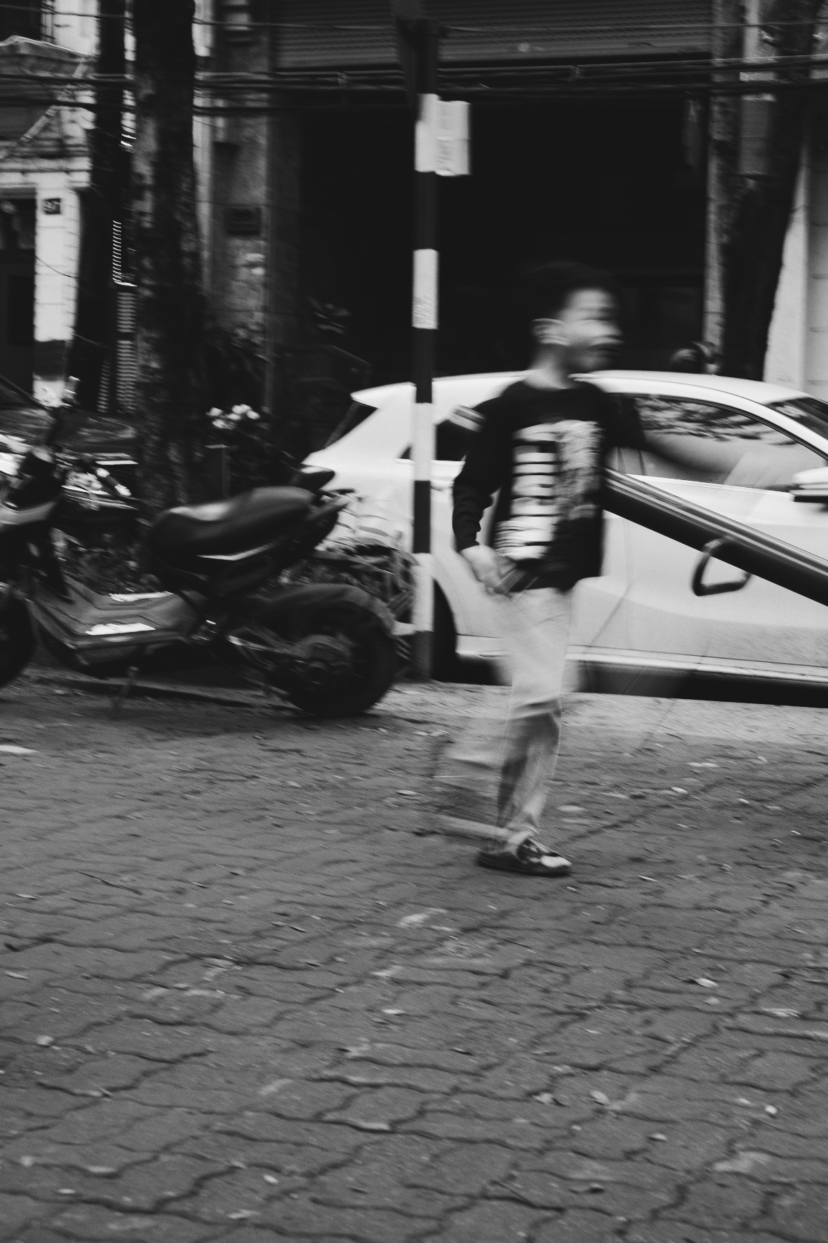 Black and white photograph of a boy playing badminton, just after hitting the ball. Blurredm He's in the middle  of a paved playground, with parking cars and motorbikes just behind.