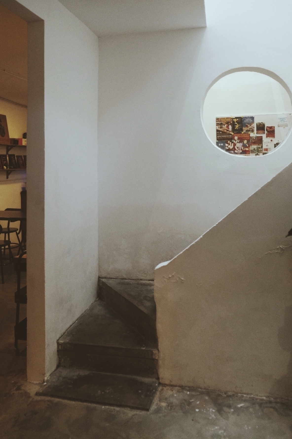 A minimalist stairway in washed white is in focus, while there's a little bit of coffee house chairs on the left in a separate room, and on the right, where the stairway goes up, there's a round peel hole with coffee decorations in sight 