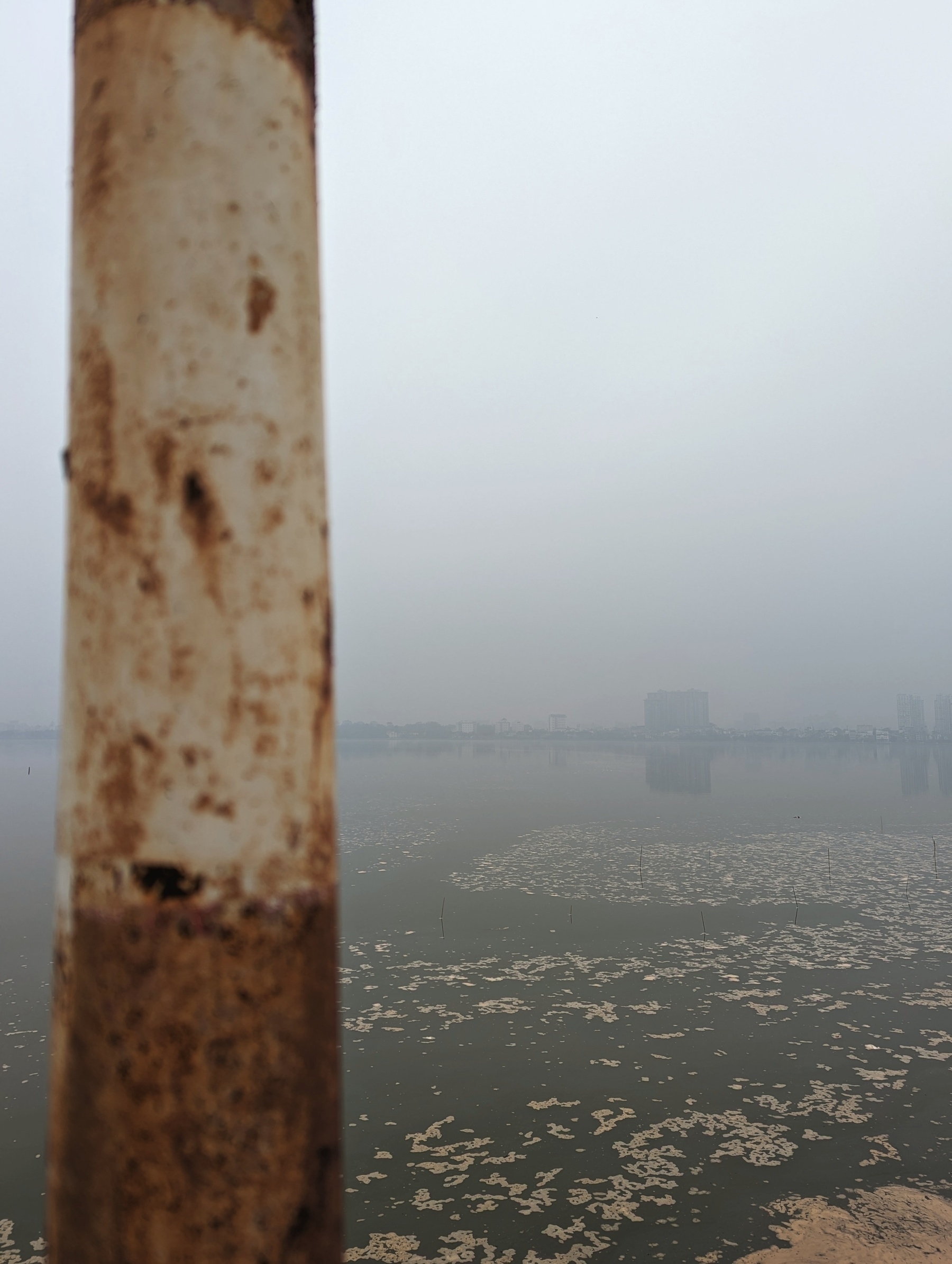 The west Lake in Hanoi in the afternoon. There's construction in the lake thus a bit of sand floating around. Most strikingly, it's foggy. A lot. Just a grey sight. There's a rusty pole on the left, because rust gives a nice aura 