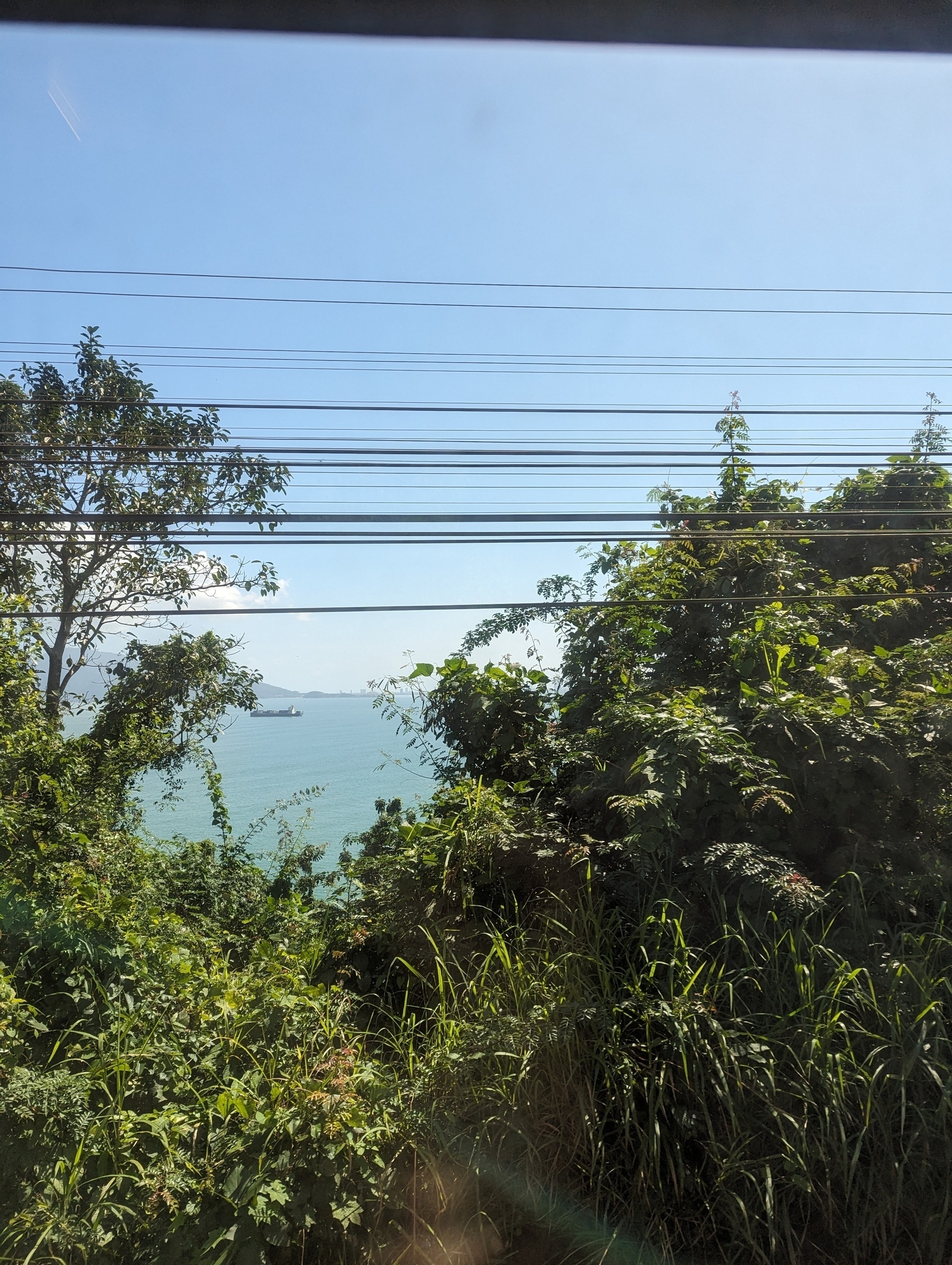 The sea shot from above through trees, or, from the railway tracks. There's a ship floating around and cables near the line 