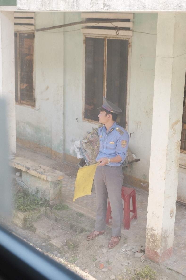 A Vietnamese train maintenance guy waiving a yellow flag, he just stood up from a red plastic chair he apparently was sitting on. He's got a nice hat 