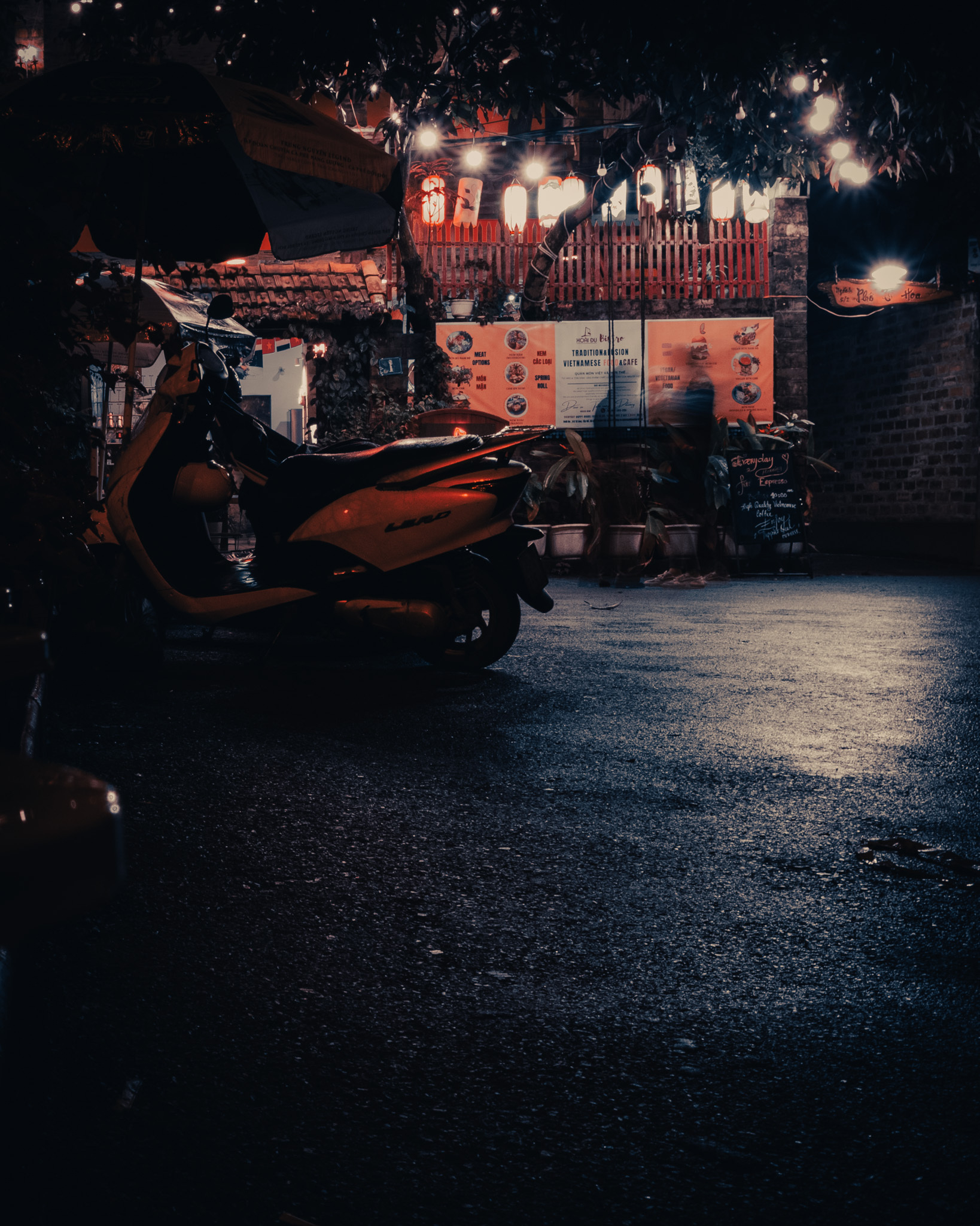 This is a dark shot with a bar/cafe glowing in orange lights in the background. Slightly hidden in focus are two persons. But these are mere shadows due to the extrem lighting/blurr. There's also a motorbike hidden in the front. Because Vietnam.